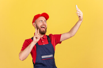 Satisfied worker in overalls taking selfie or talking on video call, looking playfully at device camera and showing v sign to client. Indoor studio shot isolated on yellow background.