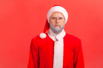 Fototapeta na wymiar Portrait of serious elderly man with gray beard wearing santa claus costume looking at camera with strict and bossy facial expression. Indoor studio shot isolated on red background.