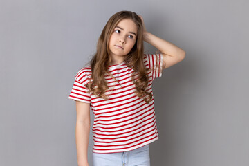 Portrait of pensive thoughtful little pretty girl wearing striped T-shirt being deeply in thoughts, keeping hand on head, looking away. Indoor studio shot isolated on gray background.