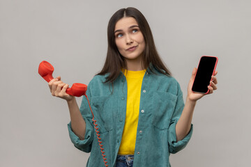 Portrait of confused puzzled woman standing retro telephone and mobile phone, thinking what to use better, wearing casual style jacket. Indoor studio shot isolated on gray background. - obrazy, fototapety, plakaty
