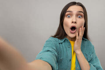 Surprised woman taking selfie with shocked facial expression, looking with big eyes and open mouth, POV, wearing casual style jacket. Indoor studio shot isolated on gray background.