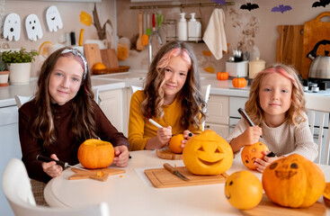 Three cute little sisters drawing and carving spooky faces on pumpkins in light white kitchen at home. Halloween party preparation.