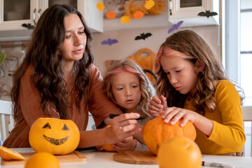 Fototapeta premium Cute young mother holding black marker to draw spooky face on pumpkin in light white kitchen at home with her happy little daughters. Halloween party preparation.