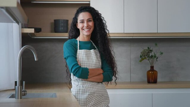 Smiling young woman maid housewife mistress baker housekeeper with long curly hair wearing checkered apron standing with folded arms confident chef posing in modern domestic kitchen cuisine interior
