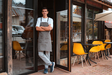 barista or waiter cafe or coffee shop owner against entrance, gesture inviting you to visit, smiling guy in apron standing outdoors being proud of his small local business