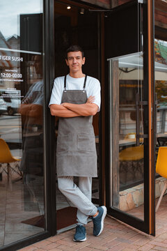 barista or waiter cafe or coffee shop owner against entrance, gesture inviting you to visit, smiling guy in apron standing outdoors being proud of his small local business