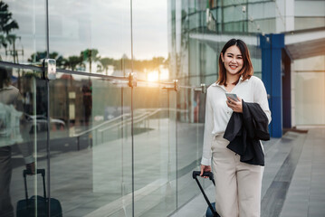 Fototapeta premium Asian woman on smart phone at gate waiting in terminal. Air travel concept with young casual business woman sitting with talking on the smartphone, carry-on hand luggage trolley.