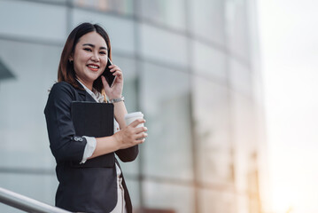 Fototapeta premium Young Asian businesswoman talking on phone and sitting in airport before business trip. Beautiful woman passenger has mobile call and discusses something with smile, holds coffee in hand