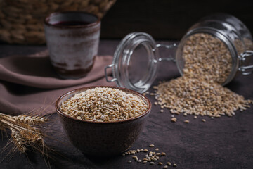 Bowl of dry raw broken pearl barley cereal grain on dark background. Cooking pearl barley porridge...
