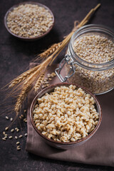 Bowl of cooked peeled barley grains porridge with ears of wheat on dark wooden background. Cooking Healthy and diet food concept.