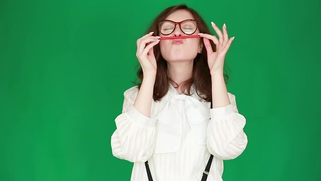 A girl in office clothes and glasses grimaces and plays with a pencil, on a green screen. Chroma Key