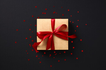 Luxury craft paper gift box with red ribbon bow with confetti stars on black background. Black Friday sale concept.
