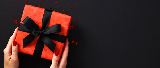 Luxury red gift box wrapped black ribbon bow in female hands over black background with confetti....