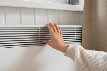 Female Hand warming over electric Heater. Frozen Woman wearing a warm woolen Sweater freezing for winter Cold. Discomfort spending time at Home. Girl's Hands on modern white Radiator. Heating problems