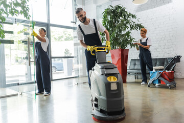 man in workwear with floor scrubber machine near interracial women cleaning office lobby.