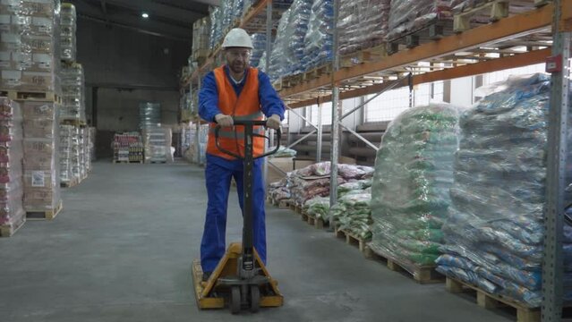 Storehouse worker having fun driving the pallet jack at the factory. Storehouse worker standing on the pallet jack. Storehouse worker bringing the pallet jack towards the delivery products.