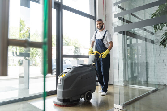 full length of professional cleaner with electrical floor scrubber machine smiling at camera.