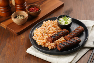Traditional south european skinless sausages cevapcici made of ground meat and spices on black plate on dark wooden board, vegan substitute made of soy protein, with rice and sour cream