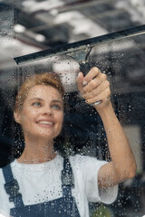 professional cleaner with window squeegee smiling near wet glass in office.