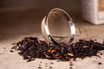 the tea strainer lies on a pile of black tea made of leaves and flowers, on a beige background,...