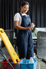 Fototapeta na wymiar cheerful mixed race woman in workwear using smartphone near cart with cleaning supplies.