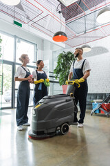 happy multiethnic professional cleaners talking near floor scrubber machine in office lobby.