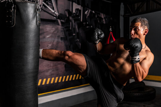 High quality photography. Shirtless muscular Hispanic man kicking a punching bag. Latin man training mixed martial arts with box gloves.