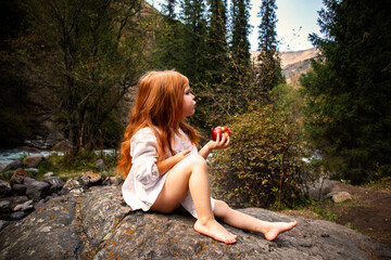 a little girl with long red hair in a white dress is sitting near the river and eating an apple