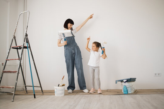 Happy family, young caucasian mother and her cute child daughter dancing and paints the wall with white paint. Repair in the apartment concept.