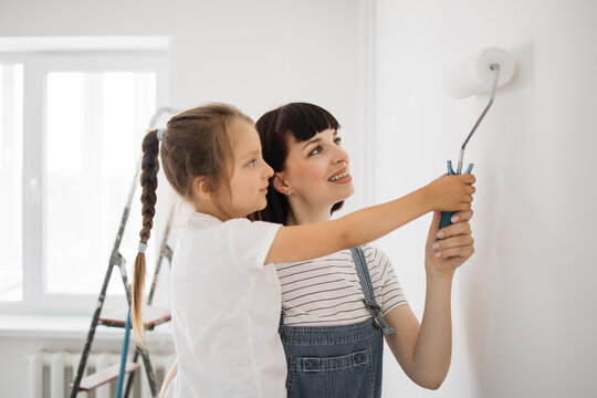 Repair in the apartment. Happy family mother and daughter paint the wall with white paint. Mother helps her daughter to paint the wall with a roller. Horizontal shoot.