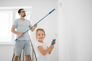 Smiling 4-year-old girl holds a paint roller in her hands. The child helps her father, who is standing behind her, near the wall, in the repair.
