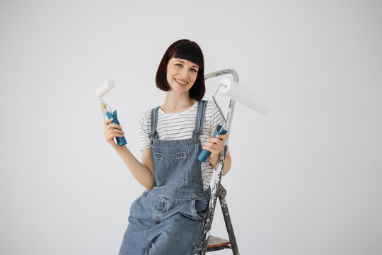 Beautiful young woman at age of 30, sitting on ladder and holding paint rollers and brush in hands with which she has just painted wall in living room.