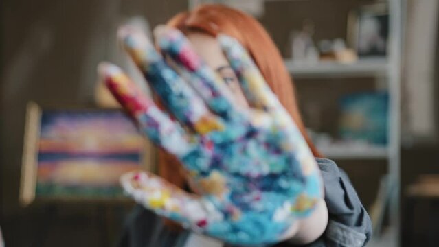 Caucasian red haired woman artist designer painter girl show dirty in paints colorful palm at camera holding hand in front of her close face with arm stop gesture not no in art studio after painting