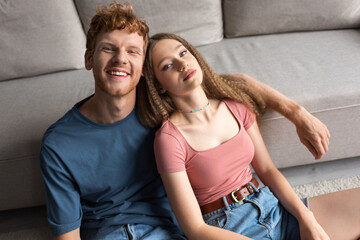 high angle view of young couple smiling while sitting near couch in modern living room.