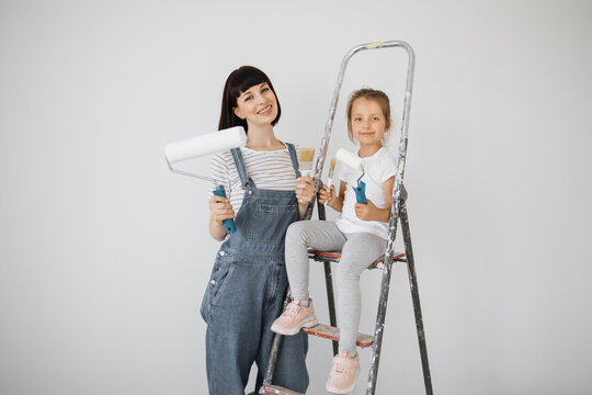 A cheerful happy family is renovating a purchased apartment. The mother hugs her daughter, sitting on a ladder, with joy. Each holds a paint roller and brushes to painting the walls.