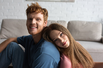 teenage girl leaning on back of cheerful boyfriend with red hair in living room.