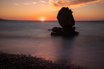 Fototapeta na wymiar Setting sun at horizon, and interesting shaped rock stands in the middle of the sea at the background in Rhodes, Greece