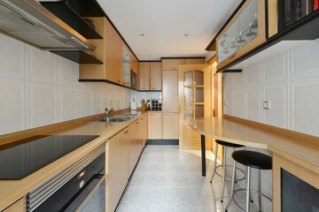kitchen furnished with beech-colored wooden furniture, table with stools on one side and kitchen...