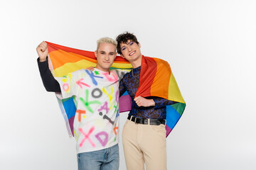 young and stylish gay man and queer person with lgbt flag smiling at camera isolated on grey.
