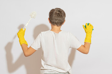 A teenager in rubber gloves for cleaning, with a plunger and a sponge in his hands, stands with his back to the camera. Against the background of a white wall.