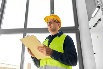 construction business and building concept - happy smiling male electrician or worker in helmet and safety west with papers on clipboard and pencil at electric board