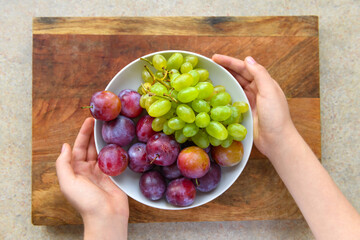 women's hands with a plate full of green grapes and plums on a wooden board, the concept of fresh fruit and healthy food