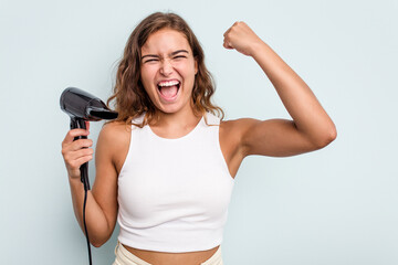Young caucasian woman holding a hairdryer isolated on blue background raising fist after a victory, winner concept.