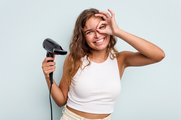 Young caucasian woman holding a hairdryer isolated on blue background excited keeping ok gesture on eye.