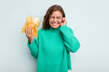 Young caucasian woman holding potato crips isolated on blue background covering ears with hands.