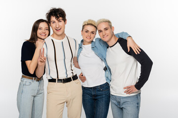 happy man showing victory sign near trendy friends hugging and looking at camera isolated on grey.