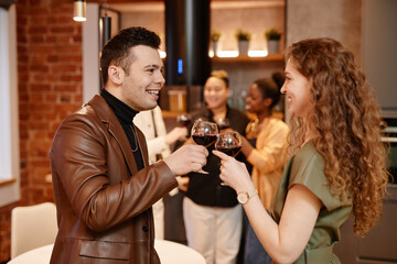 Happy young man and woman in smart casualwear clinking with wineglasses containing red wine and looking at one another