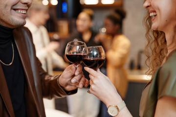 Close-up of hands of young smiling couple with red wine making toast and clinking with wineglasses afainst group of their friends