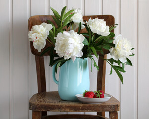 White peonies in an enameled jug and red strawberries on an old chair. Cottage core.