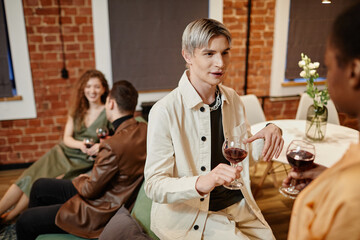 Young gay man in white casualwear holding glass of red wine and talking to African American girl standing in front of him at home party
